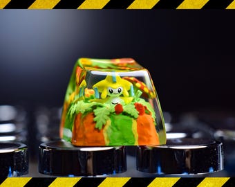 Puede incluir: Un tapón de tecla amarillo y verde con un pequeño personaje de dibujos animados amarillo en su interior. El personaje tiene una estrella en la cabeza y está rodeado de hojas verdes y zanahorias naranjas.