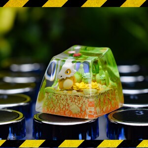 May include: A custom artisan keycap with a miniature diorama inside a clear resin enclosure. The scene includes a small, yellow and white creature, yellow flowers, and green plants. The keycap is set on a keyboard with black and silver keys.