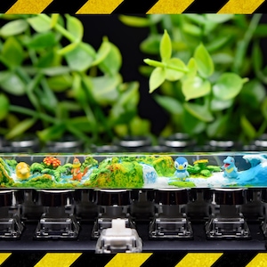 May include: A clear resin keycap with a miniature landscape scene. The scene features a waterfall, green foliage, and small blue and yellow figurines. The keycap is sitting on a black keyboard.