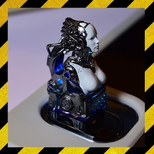 Könnte beinhalten: Eine futuristische Figur eines weiblichen Cyborgs mit weißem Gesicht und Brust sowie metallischen Silber- und Blauelementen. Die Figur weist komplizierte Details auf und steht auf einer weißen Oberfläche mit einem schwarzen, reflektierenden Sockel. Das Thema ist Science-Fiction.