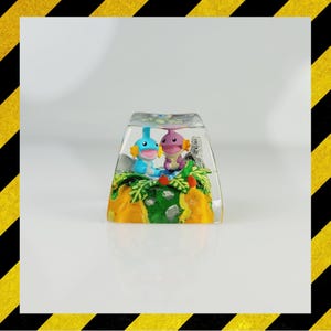May include: A clear, rectangular artisan keycap with two cartoon characters in a miniature landscape. The keycap has a yellow, green, and blue base with the characters in the center. The characters are blue and purple.