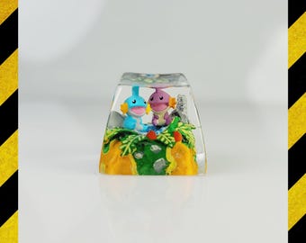 Puede incluir: Una tecla rectangular transparente con un diorama en miniatura en su interior. Dos personajes de dibujos animados, uno azul y otro morado, están situados en un paisaje con follaje verde, agua azul y una base amarilla y naranja. La tecla está sobre un fondo blanco.