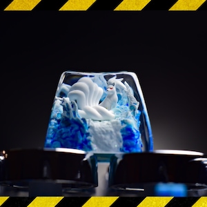 Può includere: Un cappuccio per tasti in resina trasparente con una statuetta di volpe bianca a nove code all'interno. La volpe è seduta su una base blu e bianca. Il cappuccio per tasti è circondato da altri cappucci per tasti.