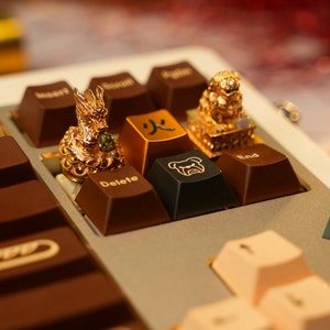 May include: A gold keyboard with brown and beige keys. The keyboard has a dragon, a lion, and a fire symbol on the keys. The keyboard is on a white surface.