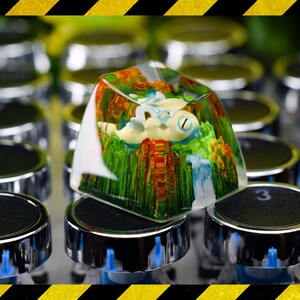 May include: A clear resin artisan keycap featuring a miniature landscape with a small white creature resting on green terrain, orange rock formations, and a blue waterfall, positioned atop a mechanical keyboard key with the number 3 visible.