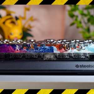 May include: A custom keyboard keycap set with a colorful, abstract design featuring a variety of Pokemon characters. The keycaps are made of clear plastic and have a glossy finish. The keycaps are mounted on a black keyboard with the SteelSeries logo.