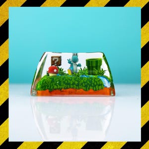 Può includere: Un keycap rettangolare trasparente con un diorama in miniatura di una scena di videogioco. La scena include un personaggio Yoshi verde, un fungo rosso, un blocco con un punto interrogativo e un tubo verde. Lo sfondo è azzurro.