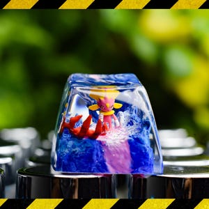 May include: A clear, rectangular keycap featuring a small figurine. The figurine is a pink and yellow character with red details, set within a blue and white water-like design. The keycap is mounted on a silver keyboard.