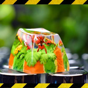 Peut inclure: Un keycap en résine transparente avec une scène miniature de créatures rouges et oranges, de feuillage vert et d'accents orange. Le keycap est posé sur un clavier, avec un arrière-plan vert flou.