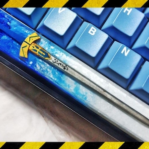Könnte beinhalten: Ein blauer und weißer Tastatur-Keycap aus Harz mit einem silbernen Schwertdesign. Das Schwert hat einen goldenen Griff und eine blaue und weiße Klinge aus Harz. Der Keycap befindet sich auf einer blauen Tastatur.