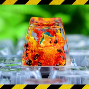May include: A clear resin mechanical keyboard keycap featuring a miniature yellow and blue creature figurine surrounded by sculpted orange, red, and yellow flames with dark grey rock-like accents embedded within the transparent casing.