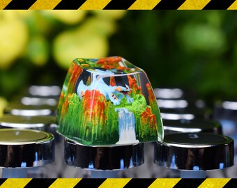 Puede incluir: Una tecla de teclado personalizada con un paisaje en miniatura que presenta un dragón azul, una cascada y follaje colorido. La tecla transparente está montada en un teclado. El diseño es visible.