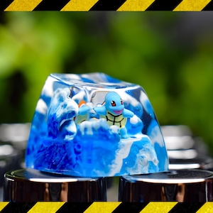 May include: A blue and white resin keycap with a small blue turtle figurine inside. The keycap is sitting on a silver surface.