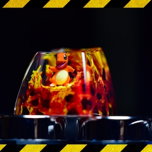 May include: A clear resin keycap with a red and orange swirl design. A small orange and yellow figurine of a Pokemon character is inside the keycap.