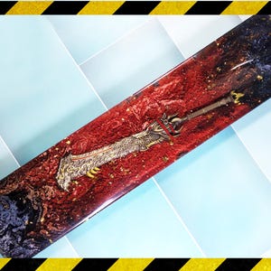Könnte beinhalten: Rechteckiger Gegenstand aus Kunstharz mit rotem und schwarzem Hintergrund, der ein detailliertes Schwertdesign aufweist. Das Schwert hat einen goldfarbenen Knauf und eine Klinge mit aufwendigen Details. Goldflitter sind im Harz verstreut.