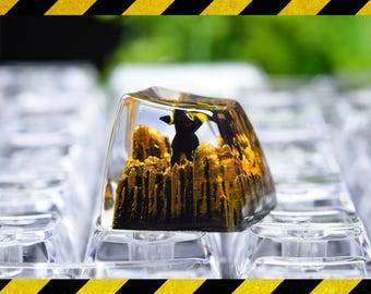 May include: A clear resin mechanical keyboard keycap featuring an embedded miniature black creature with yellow accents standing amidst a textured, golden-brown landscape that resembles a rocky terrain or forest floor, set against a dark background.