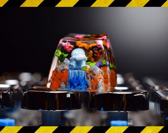 Puede incluir: Una tecla personalizada con un personaje de dibujos animados azul en una escena de cascada. La tecla transparente tiene un diseño de paisaje colorido, con elementos naranjas, verdes y rosas. Se encuentra sobre un teclado negro.