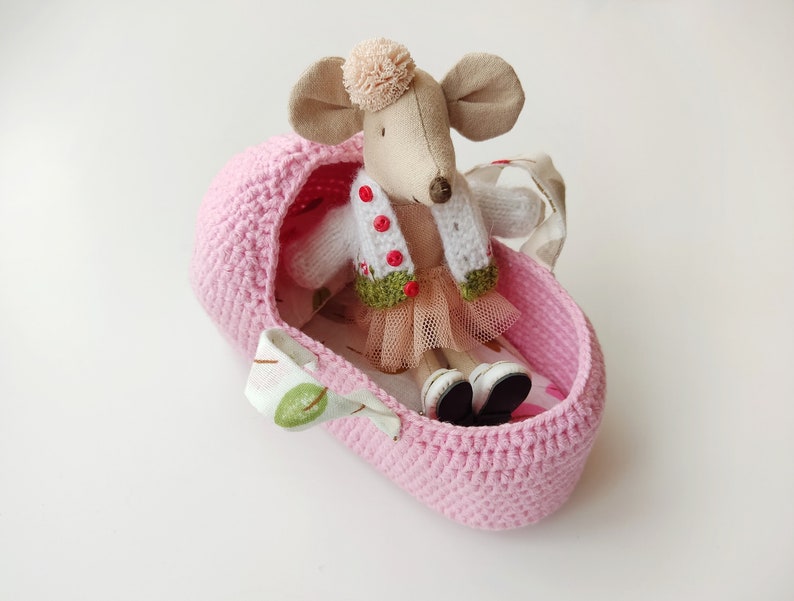 May include: A pink crocheted basket with a white and green knitted cardigan and a pink tulle skirt, holding a plush mouse toy with a brown nose and a pink pom-pom on its head.