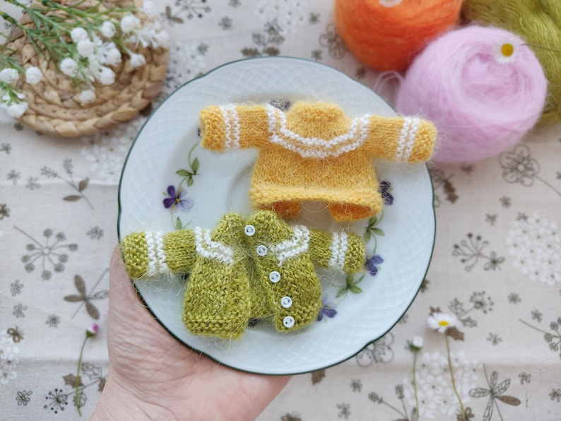 May include: Two miniature knitted sweaters, one yellow with white trim and one green with white trim, on a white plate with floral designs.