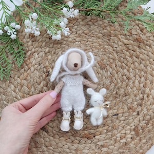 May include: Two white knitted plush toys, a bunny and a rabbit, are sitting on a woven jute mat. The bunny is wearing a white knitted hat and has a white knitted dress. The rabbit is wearing a yellow ribbon around its neck.
