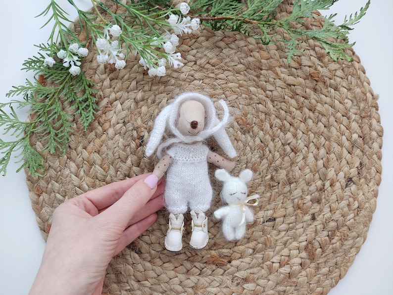 May include: Two white knitted plush toys, a bunny and a rabbit, are sitting on a woven jute mat. The bunny is wearing a white knitted hat and has a white knitted dress. The rabbit is wearing a yellow ribbon around its neck.