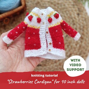 Puede incluir: Un cárdigan de punto rojo y blanco en miniatura con detalles de fresas. El suéter para muñecas presenta un cuello y puños blancos, con pequeñas fresas rojas en los hombros. El texto "Strawberries Cardigan" es visible.