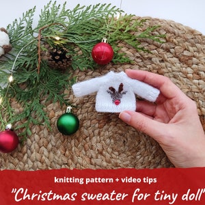 Op de afbeelding: Een kleine witte gebreide kersttrui met een rendierontwerp. De trui heeft bruine geweien en een rode neus. De afbeelding bevat de tekst "knitting pattern + video tips" en "Christmas sweater for tiny doll".