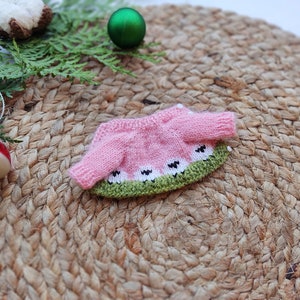 May include: A pink and green knitted sweater with a watermelon design. The sweater has white accents and is laid out on a woven surface.