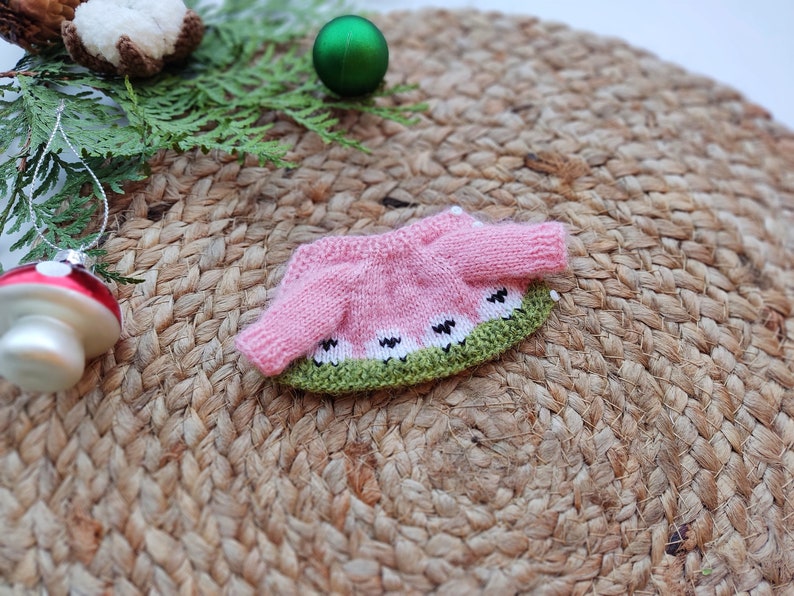 May include: A pink and green knitted sweater with a watermelon design. The sweater has white accents and is laid out on a woven surface.