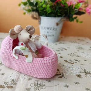 May include: A pink crocheted basket with a small stuffed mouse wearing a white dress and a red bow inside. The basket is sitting on a beige tablecloth with a floral pattern.