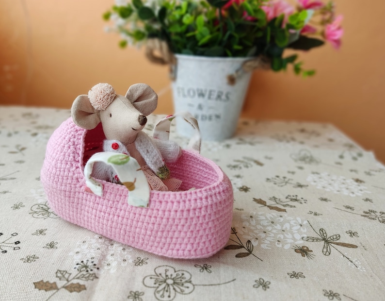 May include: A pink crocheted basket with a small stuffed mouse wearing a white dress and a red bow inside. The basket is sitting on a beige tablecloth with a floral pattern.