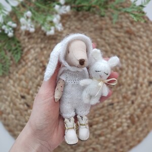 May include: A white knitted bunny doll with a white knitted hat and white boots holds a smaller white bunny doll with a pink ribbon.