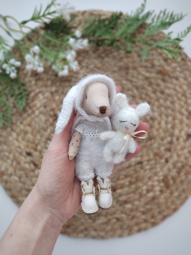 May include: A white knitted bunny doll with a white knitted hat and white boots holds a smaller white bunny doll with a pink ribbon.