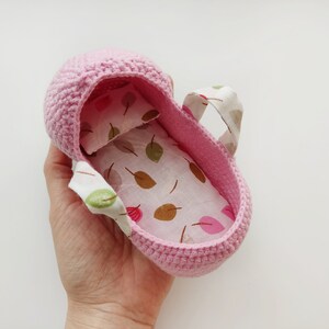 May include: A pink crocheted baby crib with a white and brown leaf patterned fabric lining. The crib has two fabric handles.