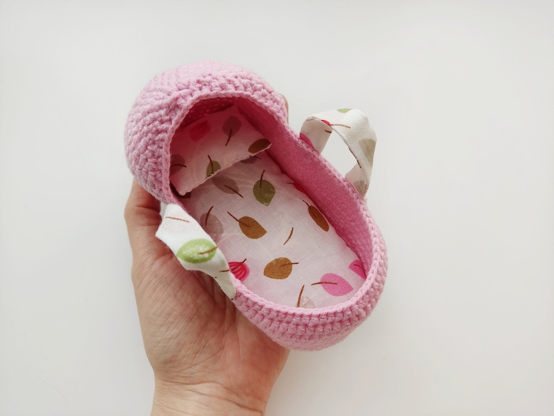 May include: A pink crocheted baby crib with a white and brown leaf patterned fabric lining. The crib has two fabric handles.