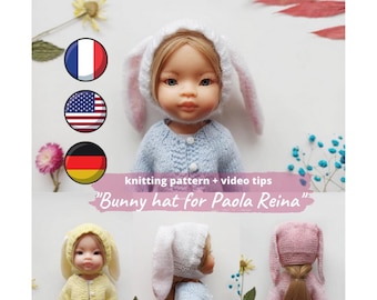 May include: A doll wearing a white bunny hat with long ears and a light blue sweater. The image includes the text "knitting pattern + video tips" and "Bunny hat for Paola Reina". Flags of France, the United States, and Germany are also visible.