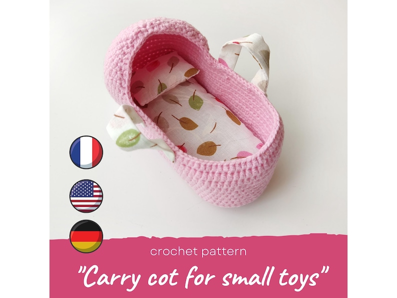 May include: Pink crocheted carry cot for small toys with a white and brown patterned fabric lining. The cot has a handle and is perfect for storing or carrying small items.
