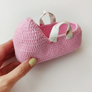 May include: A pink crocheted baby shoe with white fabric straps. The shoe is made with a delicate stitch pattern.
