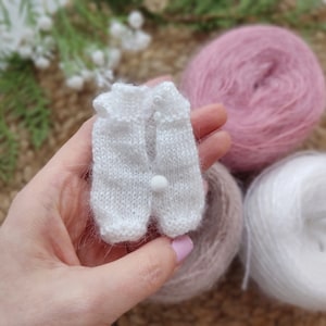 May include: A white knitted baby vest with a white button and a white pom-pom. The vest is held in a hand with pink nail polish. The vest is surrounded by balls of yarn in pink and white.