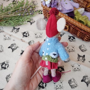 May include: A small knitted mouse doll wearing a red hat, blue sweater with white buttons, green skirt, and pink shoes. The doll is being held in a hand.