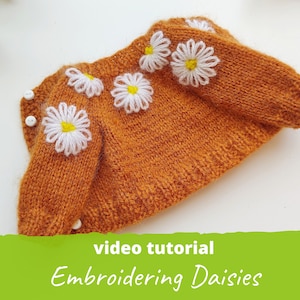 Könnte beinhalten: Nahaufnahme eines braunen Strickpullovers mit weißen gestickten Gänseblümchen. Der Pullover hat weiße Knöpfe an den Schultern. Der Text "Video-Tutorial Sticken von Gänseblümchen" befindet sich am unteren Rand des Bildes.