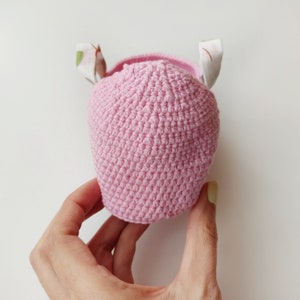 May include: A pink crocheted hat with white fabric ears. The hat is held in a hand.