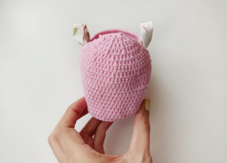 May include: A pink crocheted hat with white fabric ears. The hat is held in a hand.