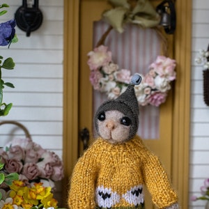 May include: A small, plush toy animal wearing a yellow jumper with a green and white pattern, a blue plaid skirt, and a grey hat with a bell. The toy is holding a basket with gardening tools and is standing in front of a miniature house with a wreath on the door.
