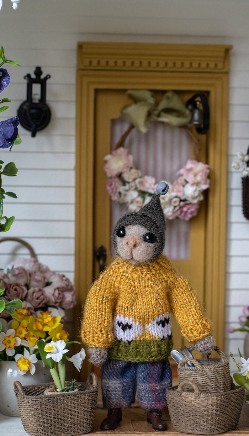 May include: A small, plush toy animal wearing a yellow jumper with a green and white pattern, a blue plaid skirt, and a grey hat with a bell. The toy is holding a basket with gardening tools and is standing in front of a miniature house with a wreath on the door.