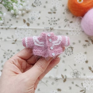 May include: A tiny pink knitted cardigan with white trim and button closures.