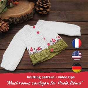 May include: White knitted baby cardigan with green and red mushroom embroidery. The cardigan has a button closure and is laid out on a wooden surface. The text "Mushrooms cardigan for Paola Reina" is visible below the cardigan.