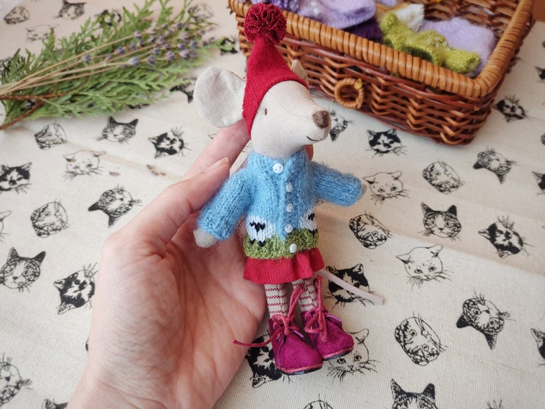 May include: A small, white mouse doll wearing a blue knitted jumper with a sheep design, a red skirt, and a red hat with a white pom-pom. The doll is holding a small, green and white knitted scarf.
