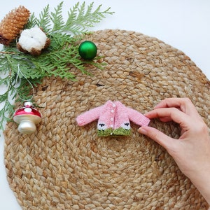 May include: A pink knitted cardigan with green trim and white buttons. The cardigan features a small sheep design on the front. The cardigan is on a woven straw mat.