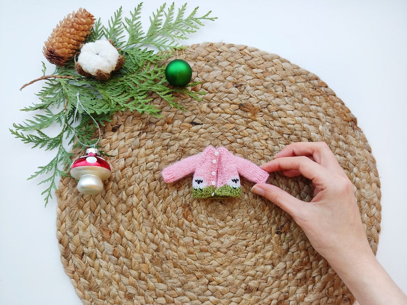 May include: A pink knitted cardigan with green trim and white buttons. The cardigan features a small sheep design on the front. The cardigan is on a woven straw mat.